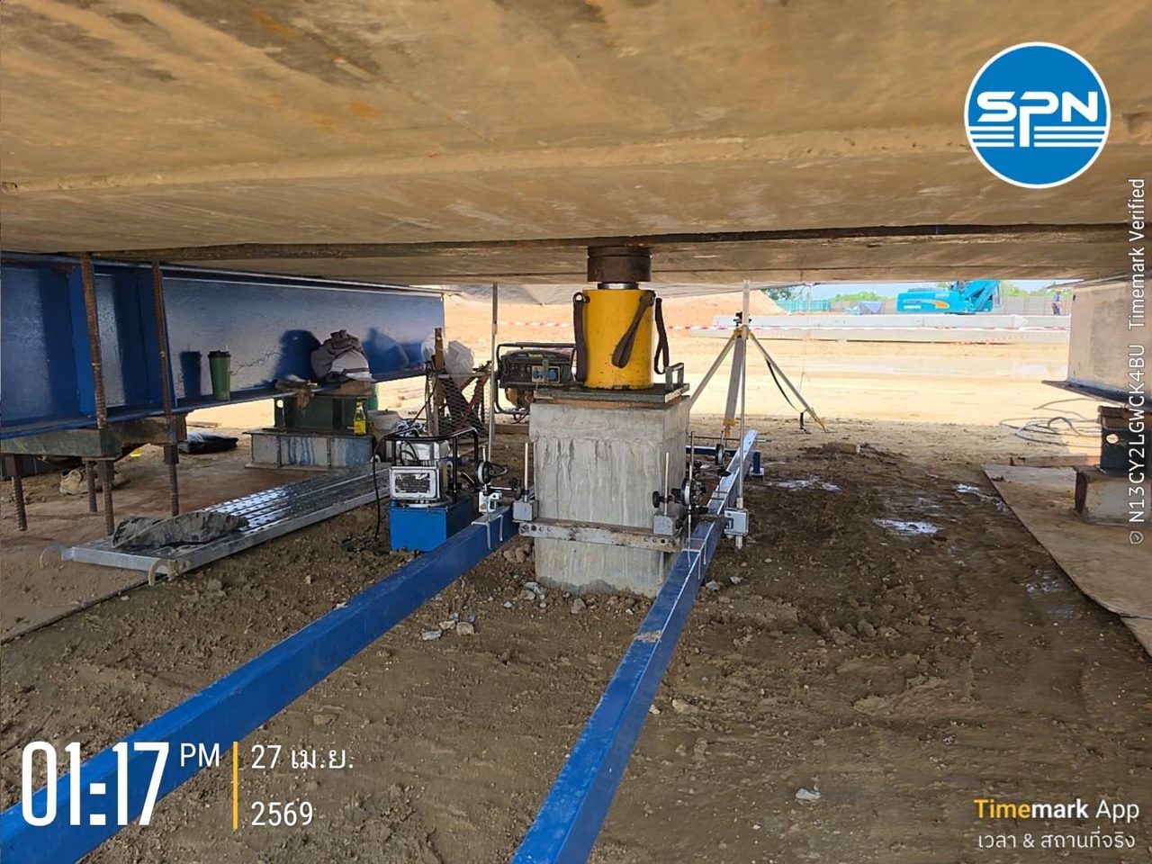 การทดสอบ Static Pile Load Test กำลังดำเนินการ — แม่แรงไฮดรอลิกบนหัวเสาเข็มใต้แท่นถ่วงน้ำหนัก (Kentledge) พร้อมมาตรวัดการทรุดตัวติดตั้งบนคานอ้างอิงอิสระสีน้ำเงิน โดยทีมวิศวกร SPN